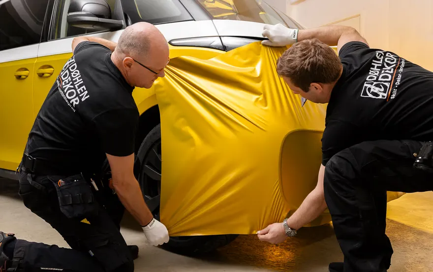 To personer som holder opp gul folie mot fronten av en VW stasjonsvogn